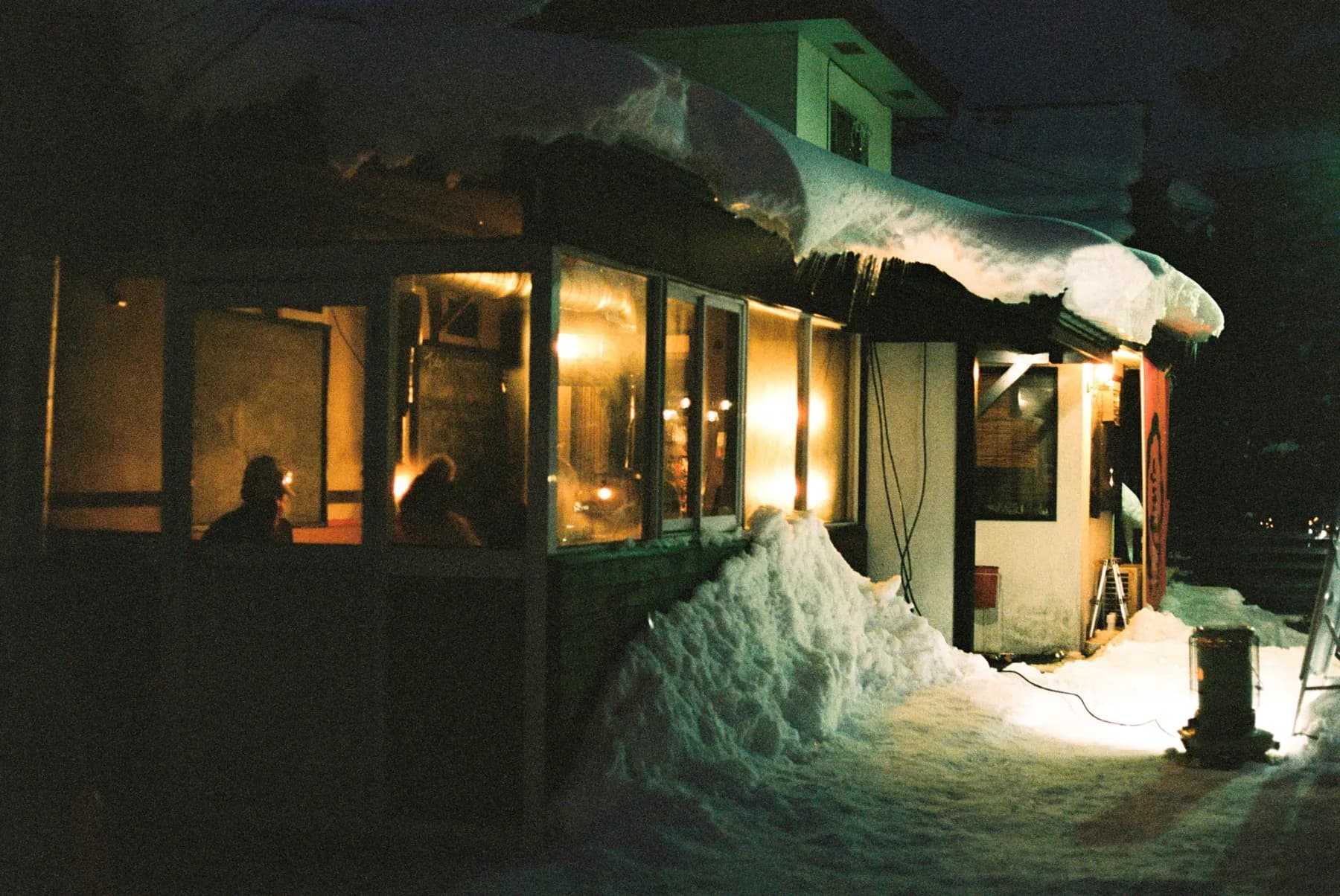 Izakaya window glowing warm between deep snowbanks at night