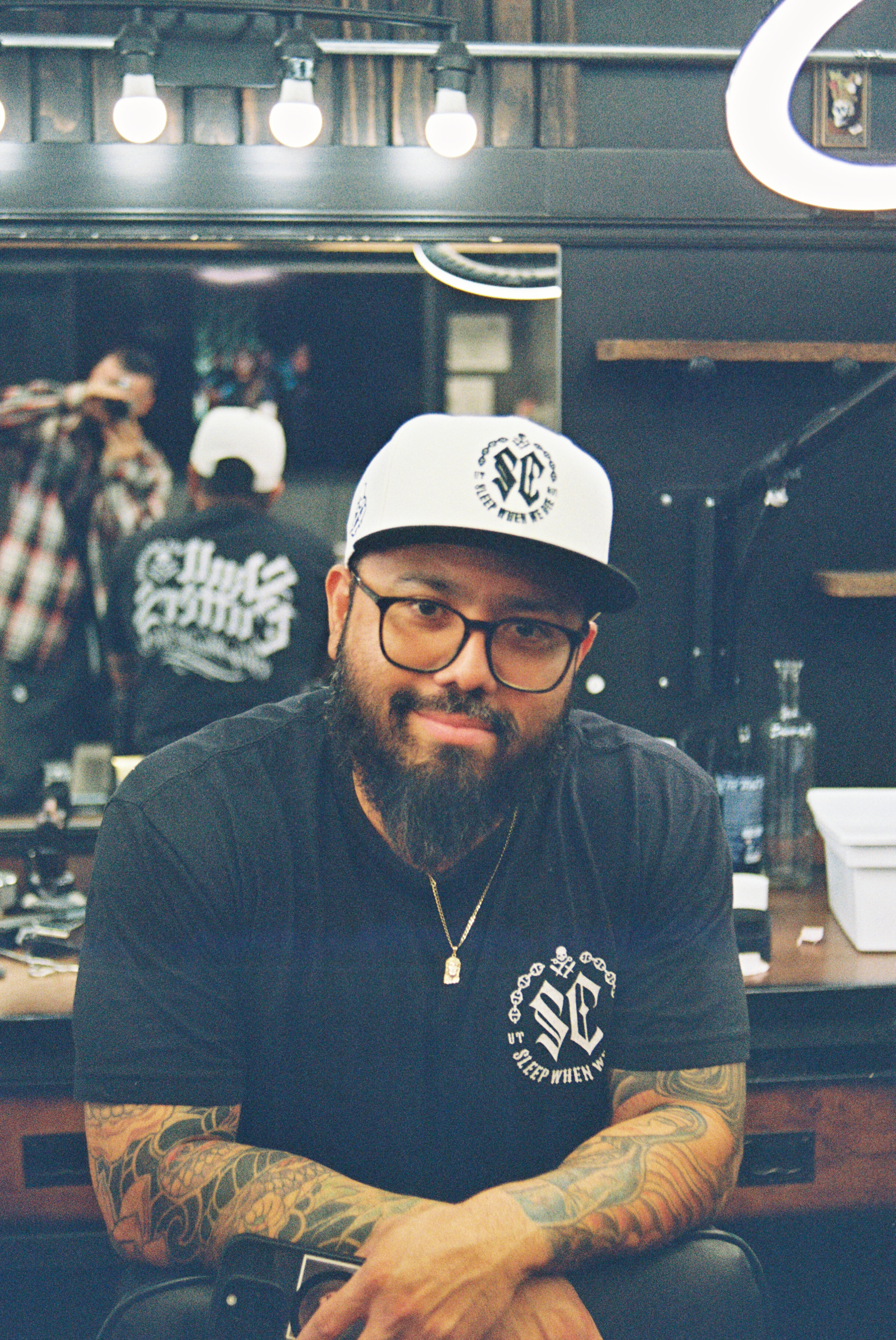 Tattooed barbershop owner in a cap and glasses, photographed on 35mm film at his chair