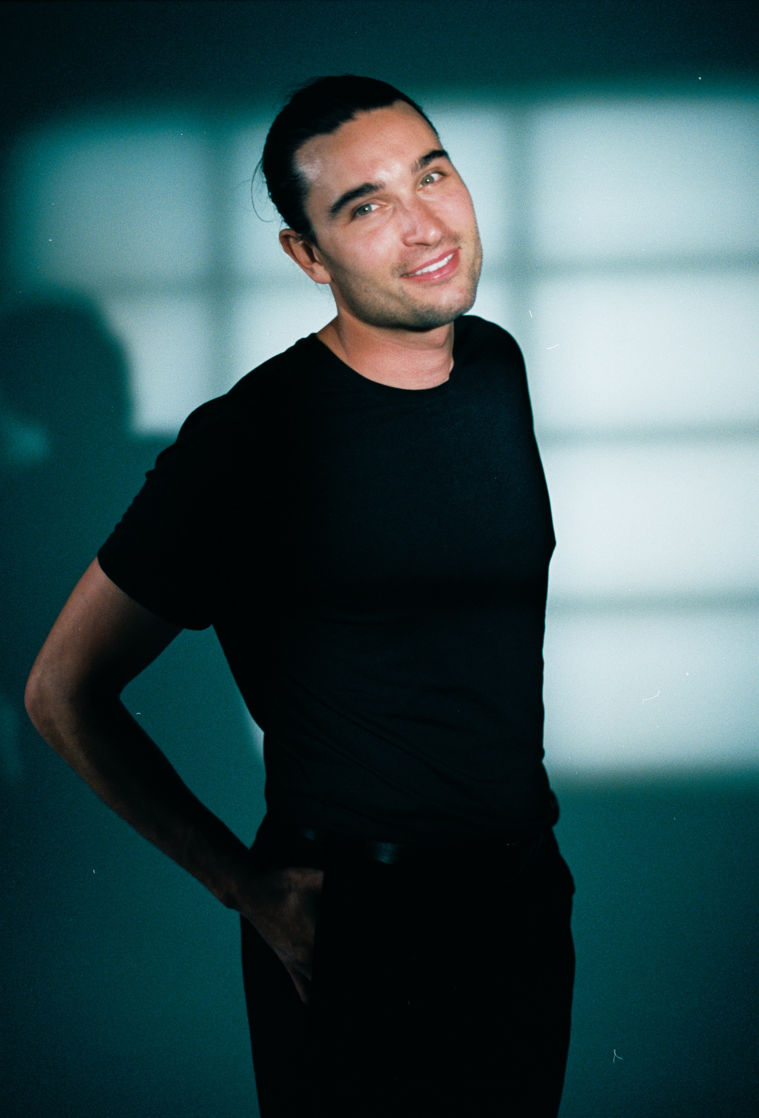 Smiling model in a black T-shirt with window-shaped light and deep teal shadows, photographed on 35mm film. cine8 cinestill
