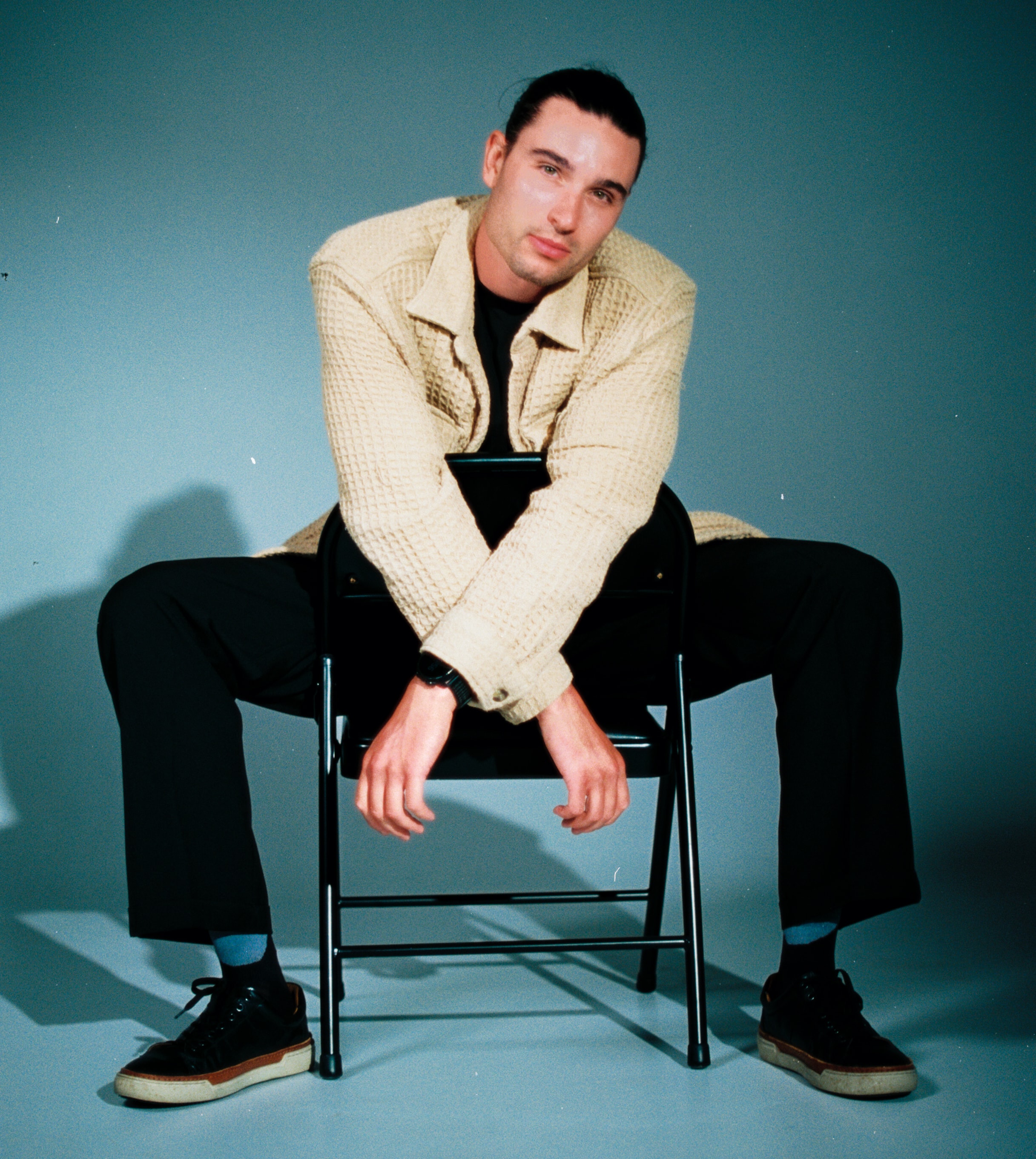 Menswear portrait on 35mm film, with a model in a textured cream overshirt sitting backward on a black chair against a blue-gray backdrop. cine 8, cinestill