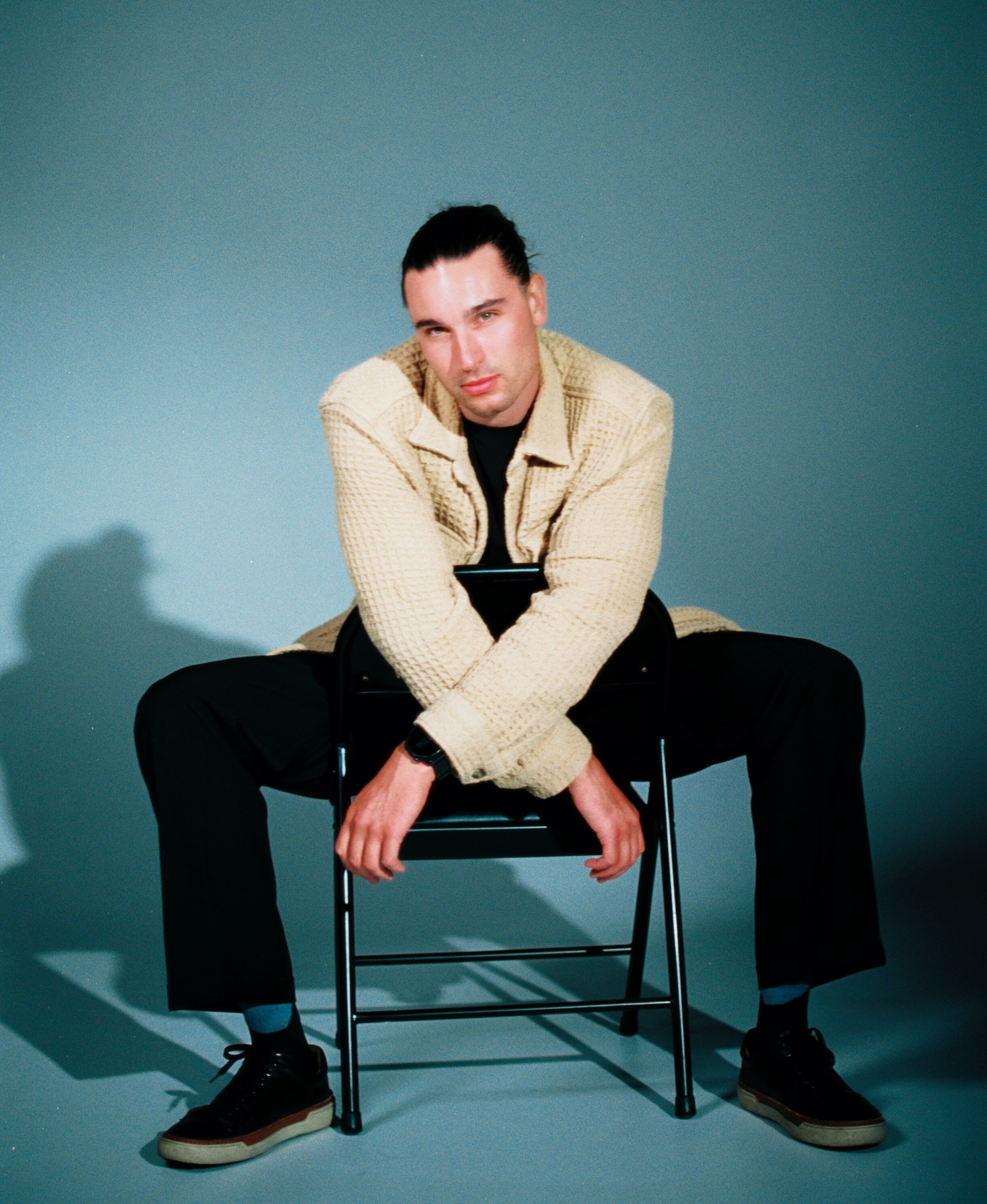 Menswear portrait on 35mm film, with a model in a textured cream overshirt sitting backward on a black chair against a blue-gray backdrop. cine 8, cinestill