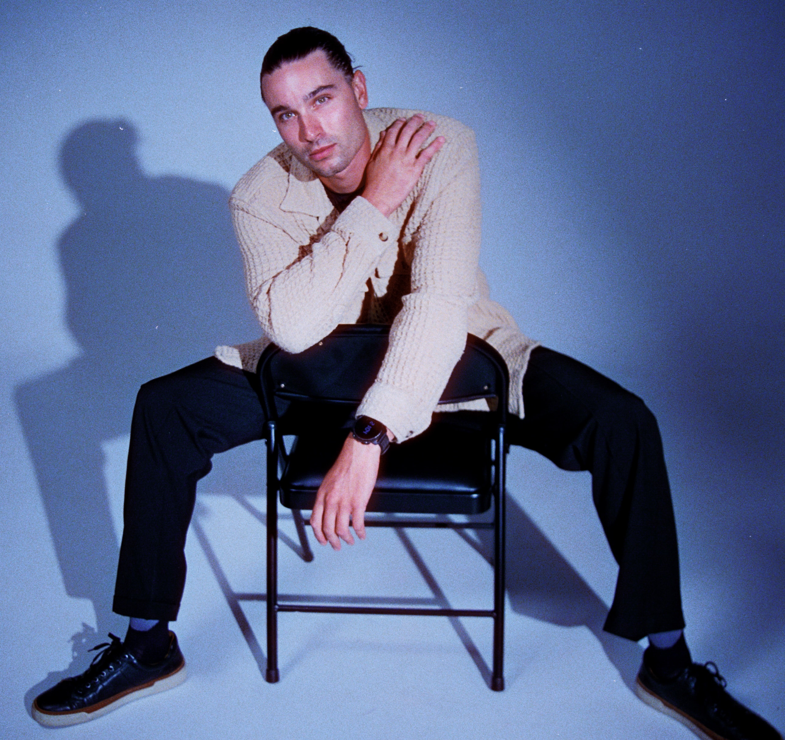 Model in a cream overshirt seated backward on a black chair, one hand resting on the opposite shoulder, with a strong studio shadow. 35 mm film, AE1
