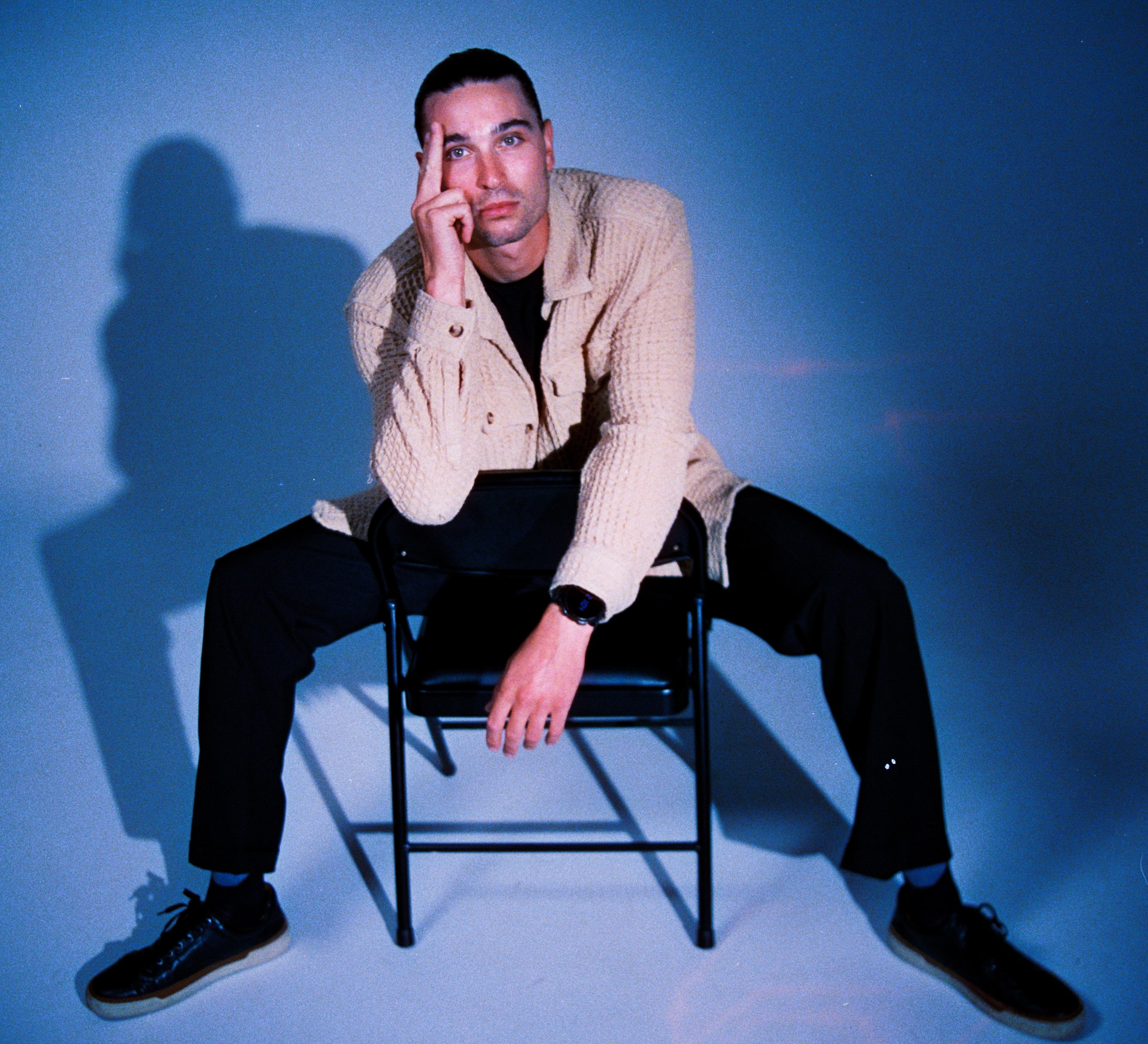 Editorial film portrait of a model leaning over a black chair with a finger against the temple, framed by blue light and a deep shadow.